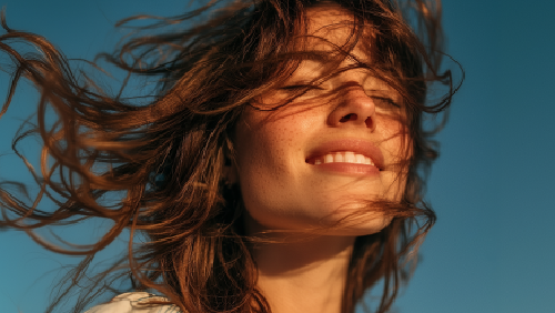 Pessoa em primeiro plano ao ar livre com o cabelo movimentado pelo vento, sob luz natural, com céu azul ao fundo, associada a conceito de leveza ou tranquilidade.