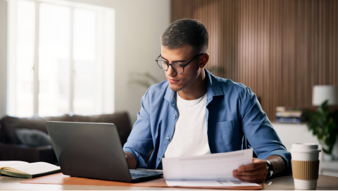 Homem jovem ao computador com documentos sobre certificados de aforro e do tesouro.