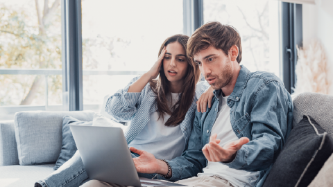 Duas pessoas sentadas num sofá a olhar para um computador portátil, com ambas as mãos gesticulando durante a utilização, em ambiente doméstico interior com janela ao fundo.