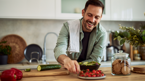 Pessoa a cortar tomate‑cereja sobre uma tábua de madeira enquanto prepara uma salada numa cozinha, com pepino cortado e um frasco de bolachas ao lado.